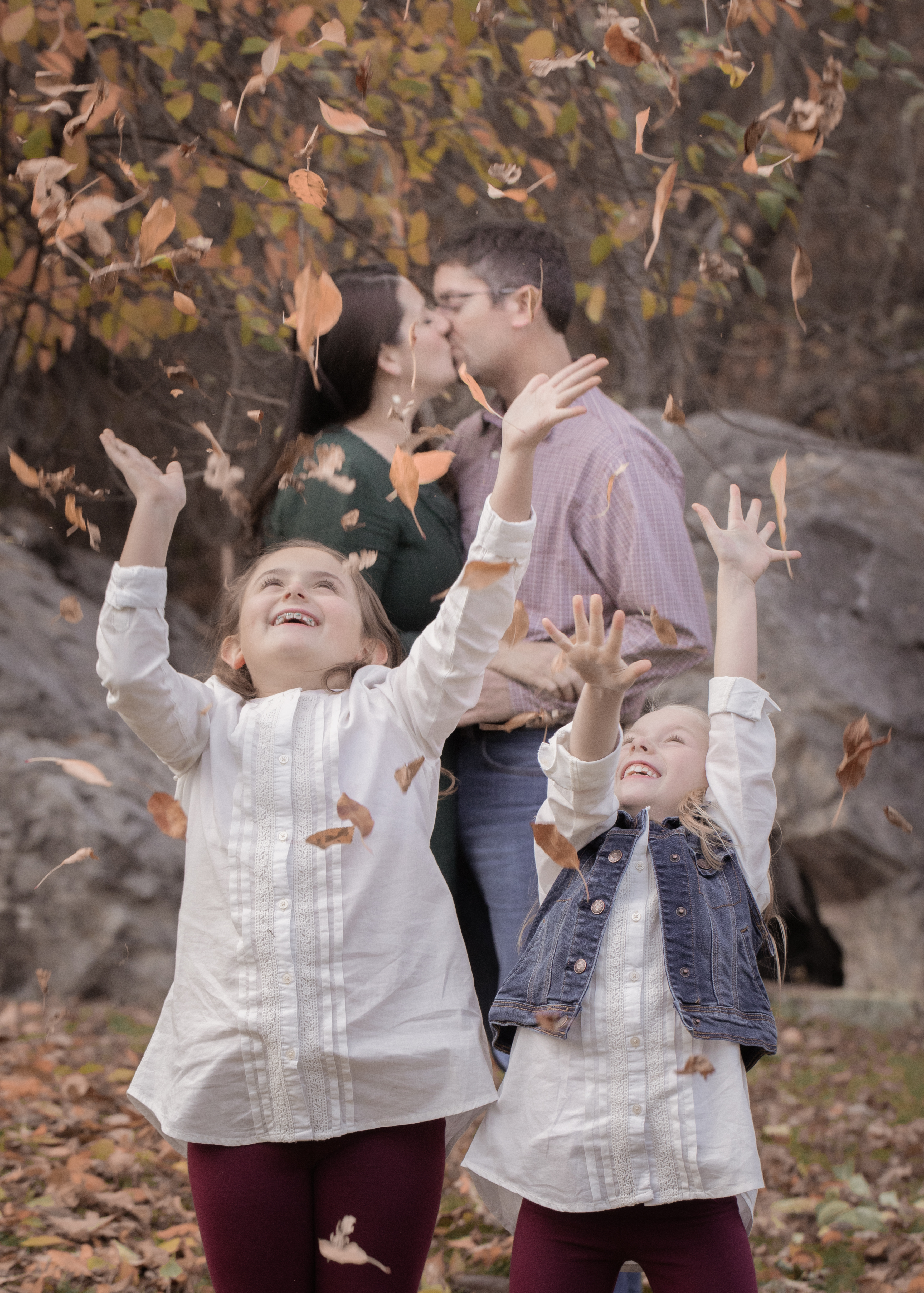 Two sisters throw dry leaves in the air while mom and dad steal a kiss in the background.